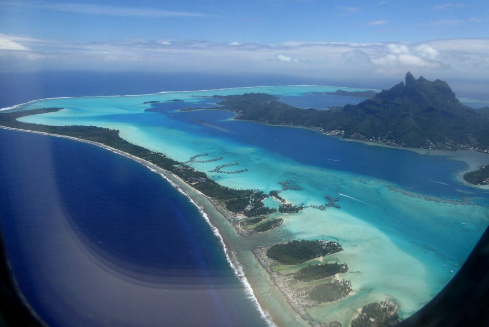 Vue panoramique de Bora Bora depuis le hublot de l'avion : motus avec bungalows sur pilotis, lagon aux nuances de bleu turquoise et mont Otemanu, Polynésie française