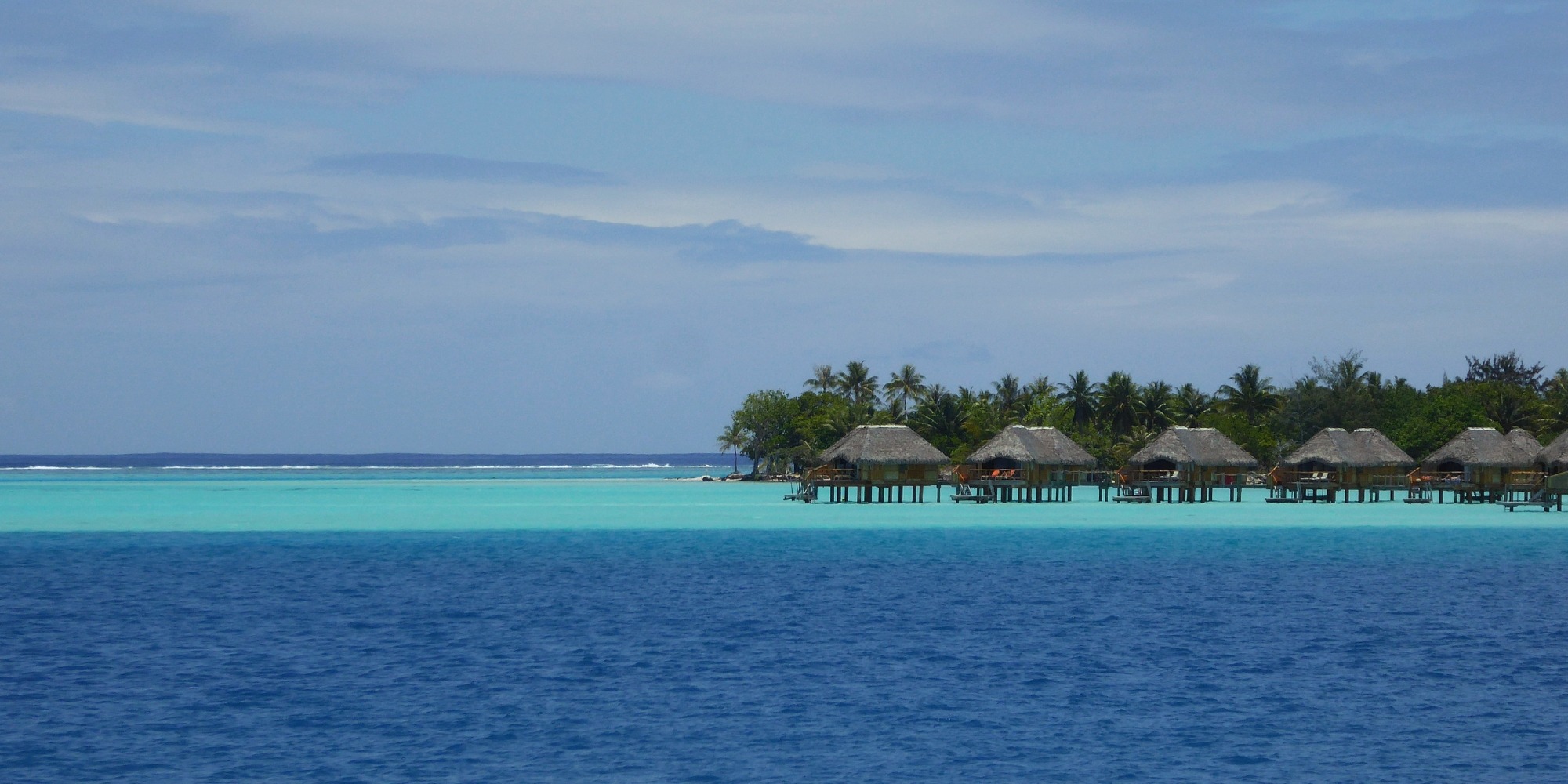 Bungalows sur pilotis sur un motu — lagon bleu de Bora Bora, Polynésie française Rangée de bungalows sur pilotis au toit de chaume sur un motu bordé de cocotiers, lagon aux nuances de bleu profond et turquoise, Bora Bora, Polynésie française