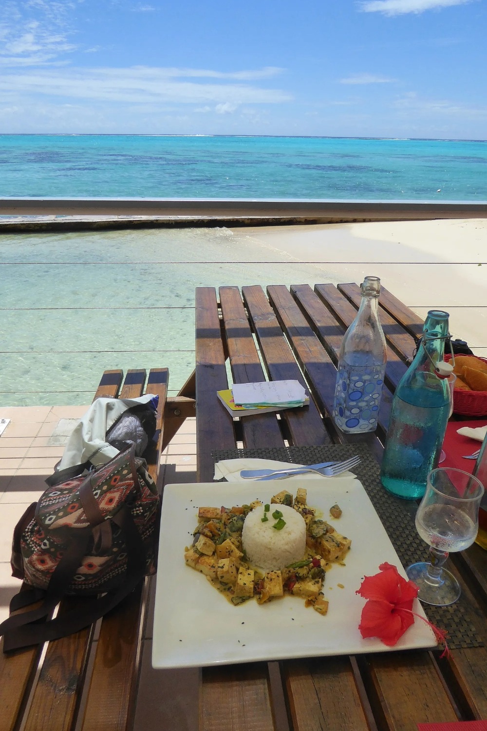 Assiette de riz et légumes polynésiens avec hibiscus rouge sur une table en bois face au lagon turquoise de Moorea depuis un restaurant en bord de mer, Polynésie française