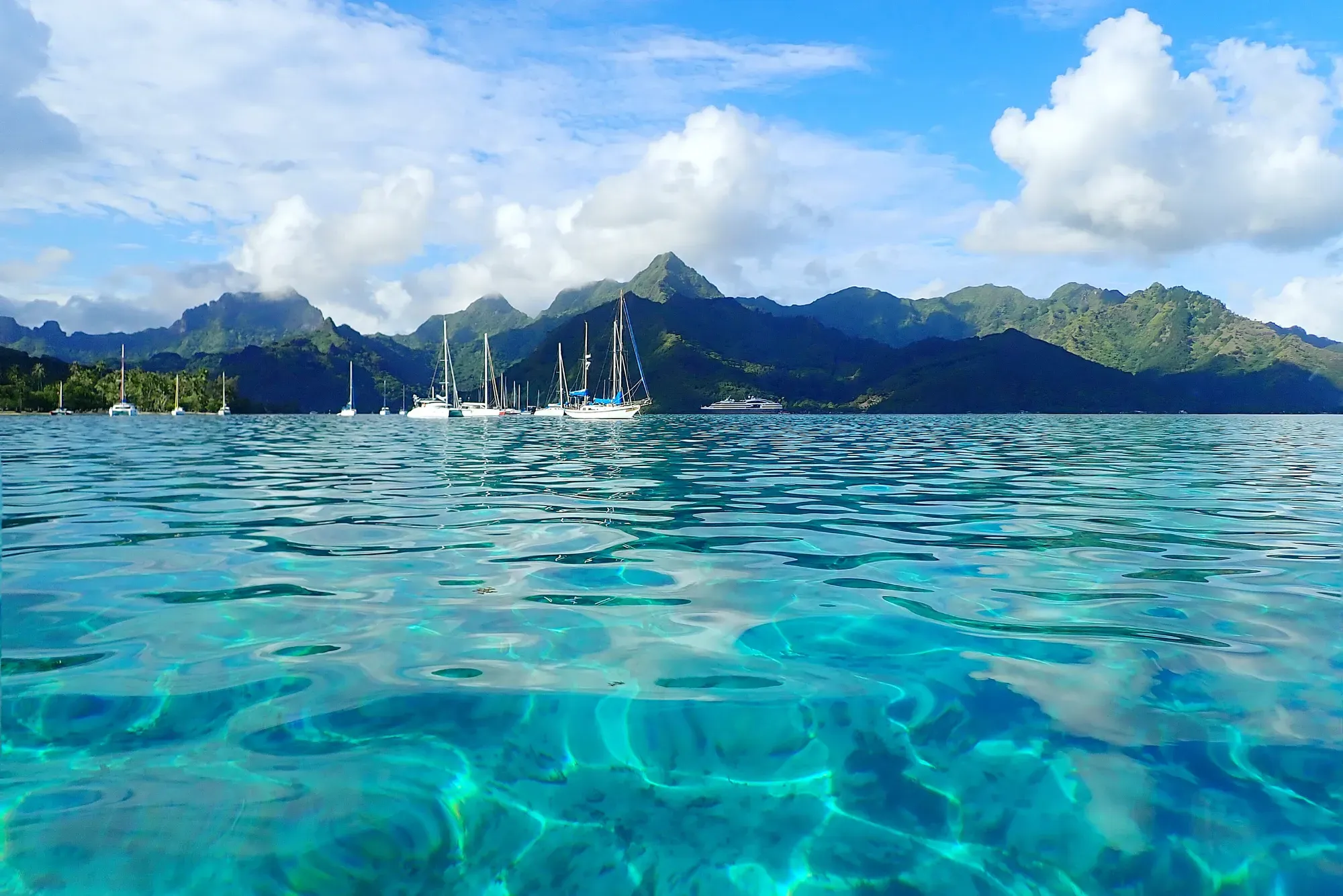 Lagon turquoise de Moorea avec voiliers et montagnes — Polynésie française Lagon turquoise cristallin de Moorea avec voiliers ancrés et montagnes volcaniques verdoyantes en arrière-plan, photographié au ras de l'eau, Polynésie française
